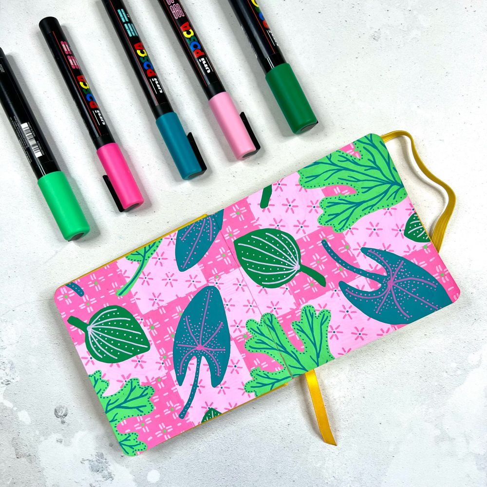 Photograph of an open sketchbook. On the open page is a repeat pattern consisting of a pink checkerboard pattern of light and medium punk squares with sashiko pattern print on them. Over the top are leaves.
There are three different shaped leaves each a different shade of green. Above the sketchbook are posca pens.