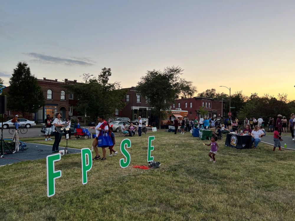 A crowd of people of varying ages stands around in a public park. Large green letters that spell out FPSE. Children play, and community organizations sit at booths talking to people.