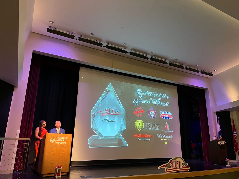 A stage with two people, Greg FX Daly, a white man wearing a blue suit, and Shameem Clark Hubbard, a black woman wearing an orange dress, both standing at a podium. Next to them, a large screen shows the picture of an award shaped like a jewel, and the text advertises “The 2021 and 2022 Jewel Awards” along with many sponsors of the event. At the front of the stage, the STLTV logo sits. The logo is the letters STL in red, TV in black, with a sweeping stylized arch over the letters and a flowing red line underneath.