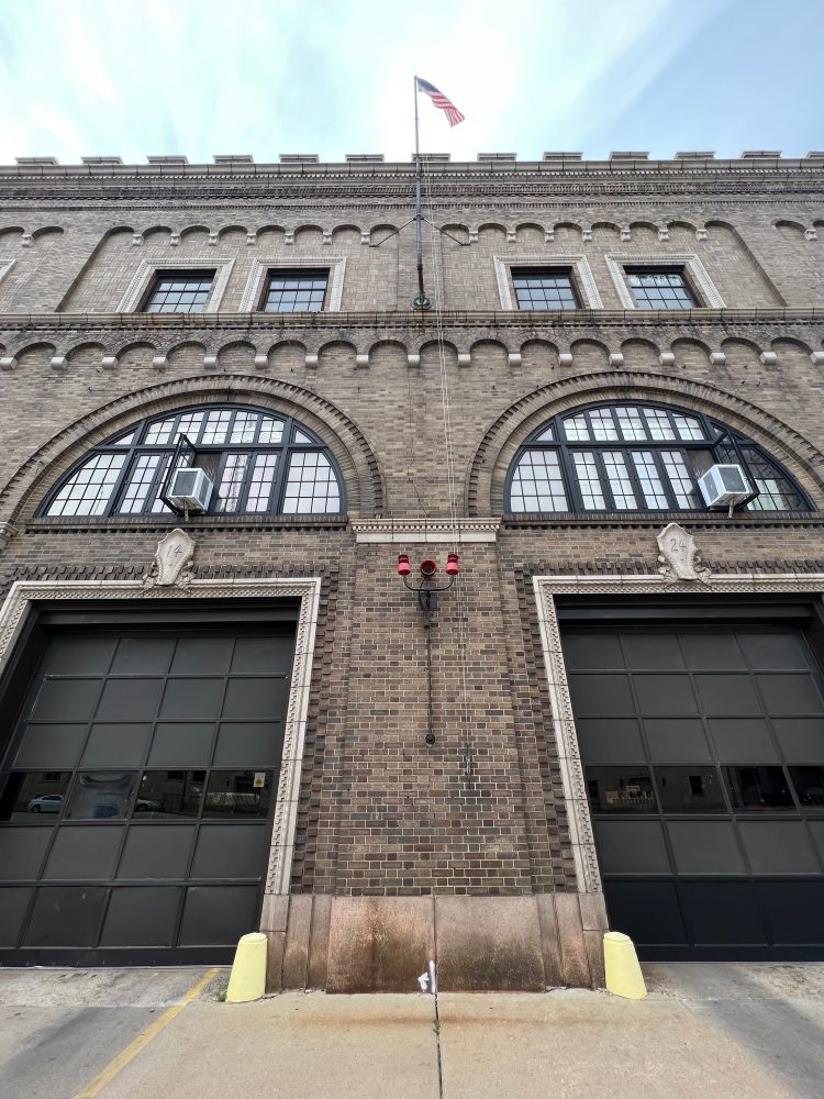 A view of the old downtown firehouse at 314 Tucker. It is built of old brown and black brick. In this picture, you can see two big bay doors where the fire trucks exit. There are two red lights in the middle between the doors, that flash to warn a truck is coming out. Above, two half circle windows sit above the doors. The top of the building has crenellations, like a castle. 
