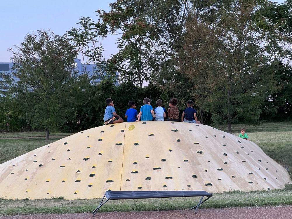 Six kids sit on top of a climbing wall on the side of a yellow mound in a public park. A seventh child climbs up the side to join them.