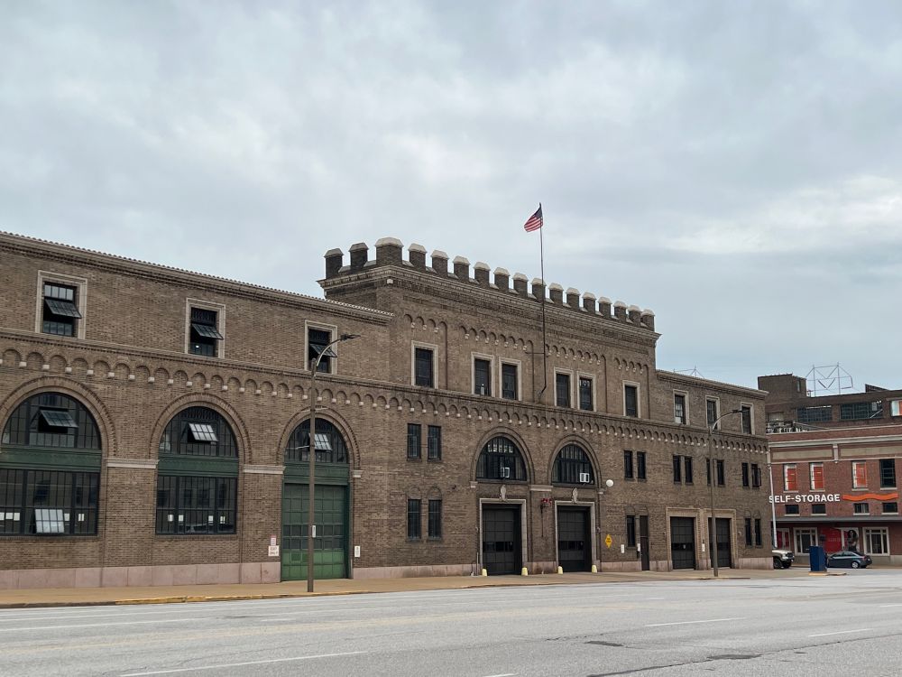 A wide shot of the whole downtown firehouse. It is an old brick building with crenelations at the top of the center of the building. Large doors and windows on the front of the building feature half-circle windows above. A lone American flag flies atop the building. 