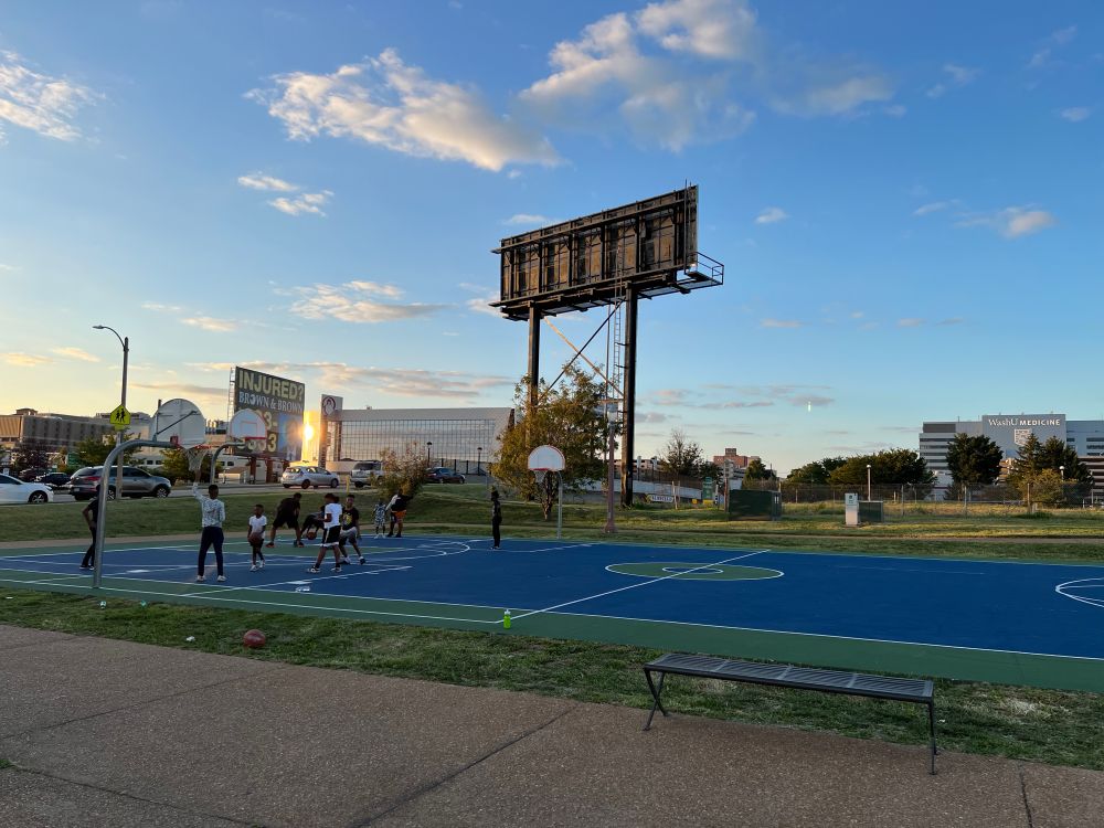 Kids of varying ages play basketball on a blue basketball court underneath a billboard. The sun sets behind them.