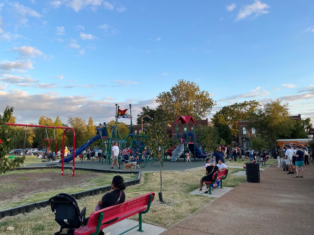 People wait in line at a food truck, while some others sit on a bench while children play on playground equipment.