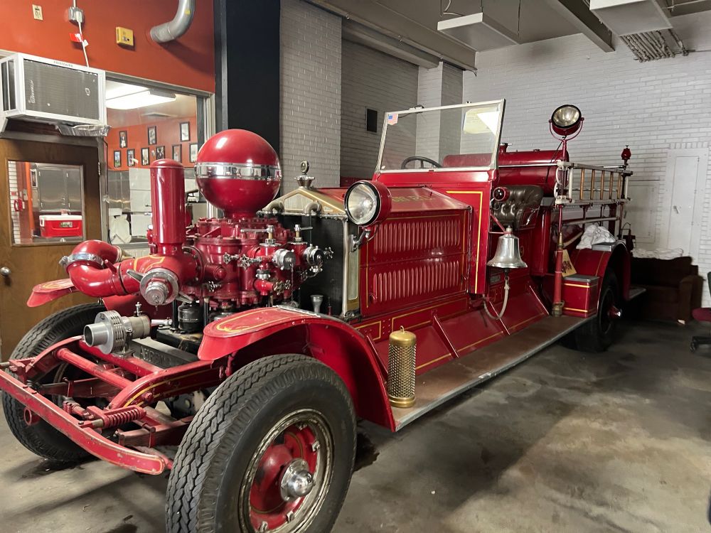 An old-fashion fire truck. It is red, has the front tires all the way up front, features a small ladder on the side, and a big silver colored bell to alert folks that it’s coming. 