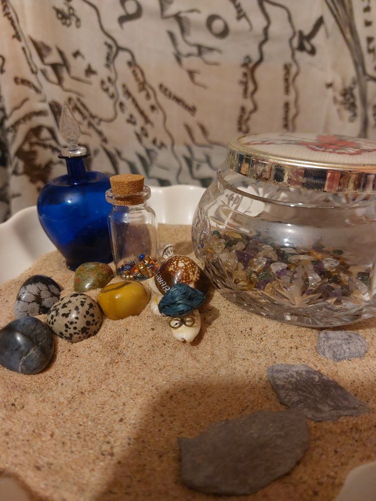 A map of Tolkien's Middle Earth hangs behind a bowl of sand. On the sand a bottle of bright beads sit next to a crystal jar of semi precious stones and a blue stopped second bottle. Semi precious polished stone eggs are guarded by a bespectacled turtle made from shells and stones. A flint pathway leads on