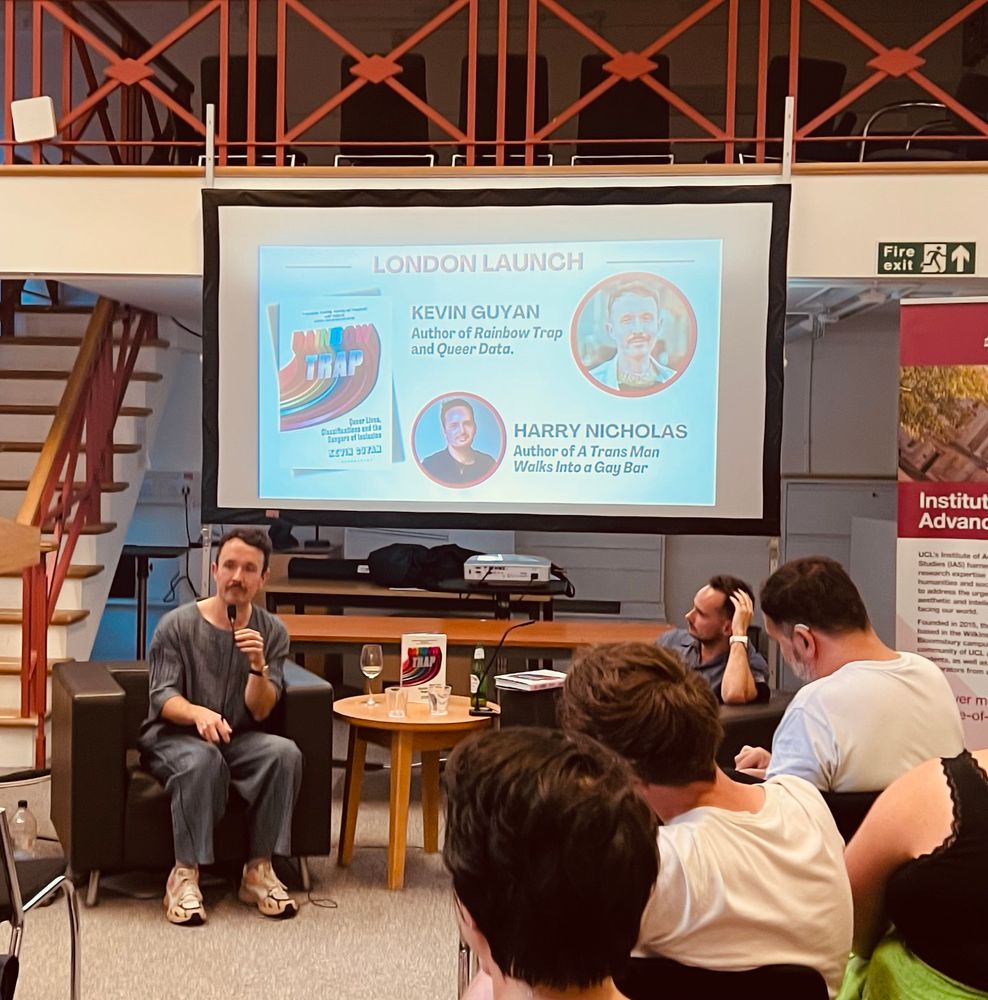 Two people sitting at table in front of a large screen highlighting London Launch of Kevin Guyan’s new book ‘Rainbow Trap’. Heads of other people are in the foreground 
