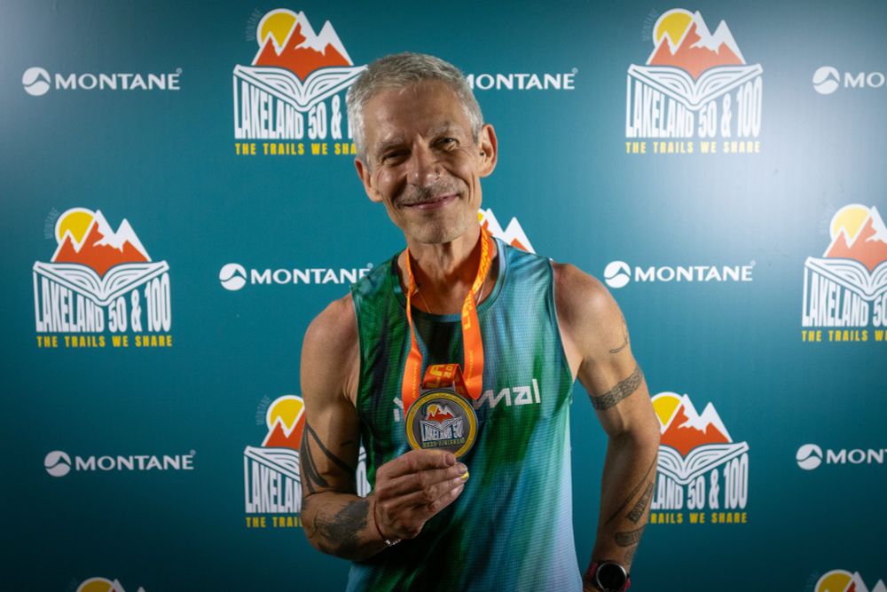 Anson in a teal coloured vest holding a finishers medal for Lakeland50. They are standing in front of a teal blue backdrop with a mountain logo and text saying Lakeland 50 & 100, and Montane 
