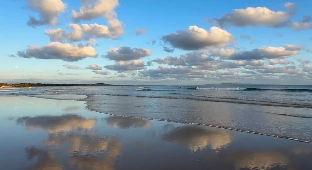 Fluffy clouds against a blue sky reflected in wet sand on the beach with gentle waves