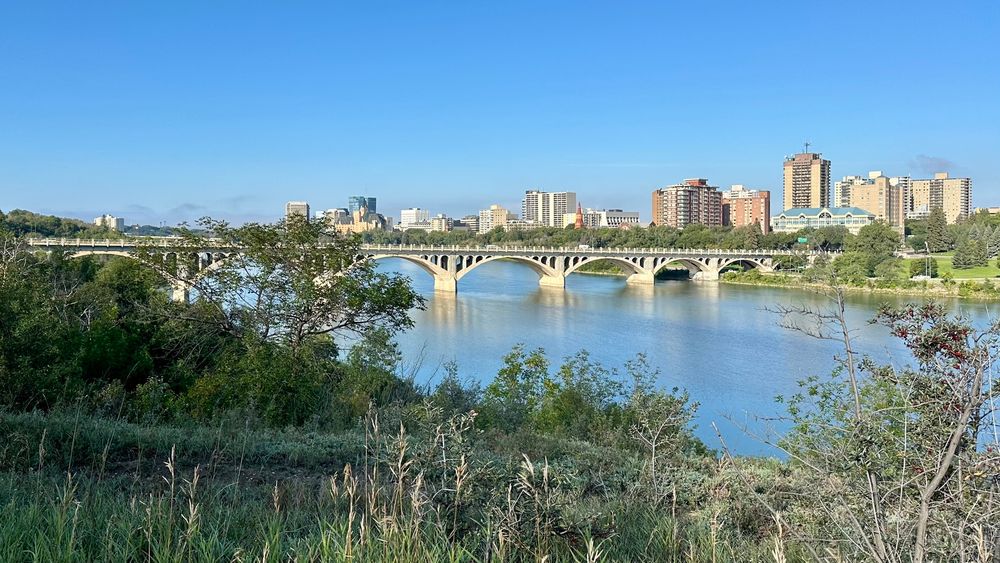 Arched concrete bridge over a prairie river with city skyline beyond. 