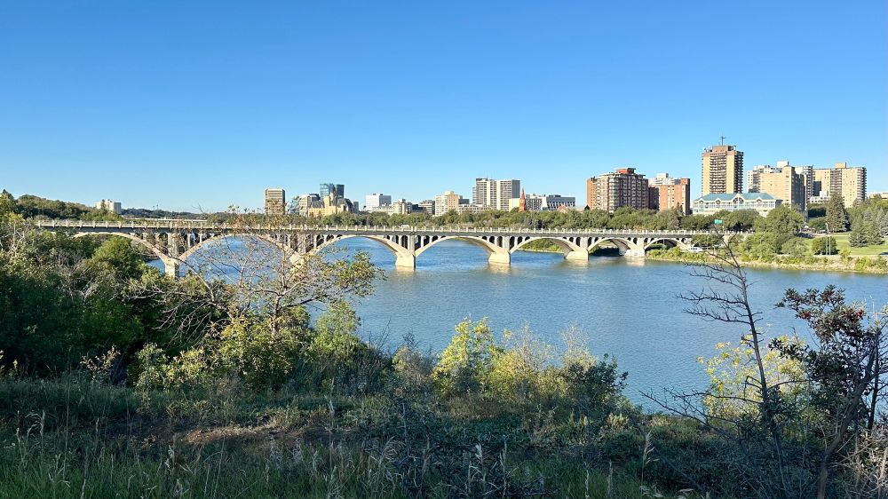 Arched concrete bridge across a prairie river with city skyline beyond. 