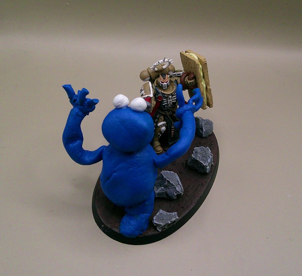 A chaplain miniature with an assault trooper head and his crozius replaced with an MDF / green stuff shield in the design of a custard cream. He is shooting at the Cookie Monster who is also made of green stuff.