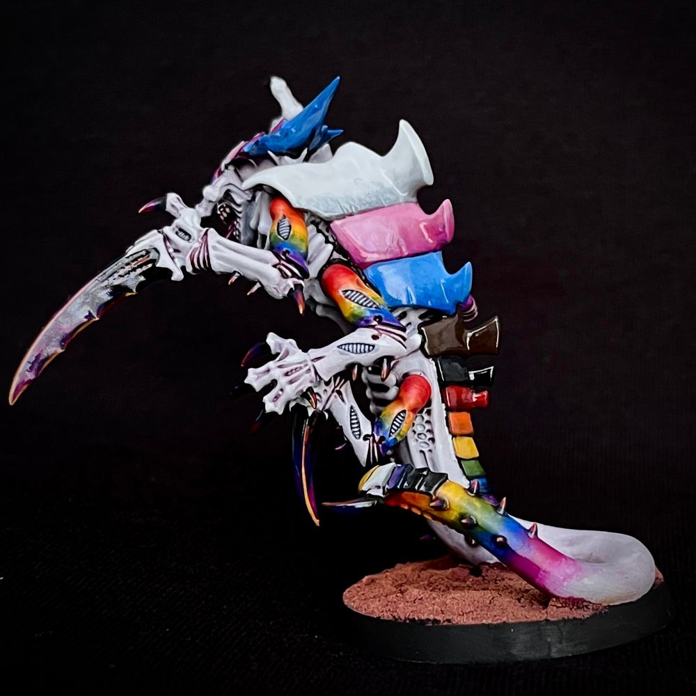 A Tyranid Ravener painted with whiteish purplish skin. Its upper arms and tail end are rainbow-hued, and its claws are purple.
It’s carapace plates are each painted a solid colour: the head matches the trans pride flag, the chest matches the bisexual pride flag, the tail matches the non-binary flag and the back matches the progress pride flag.
This Tyranid eats fascists.