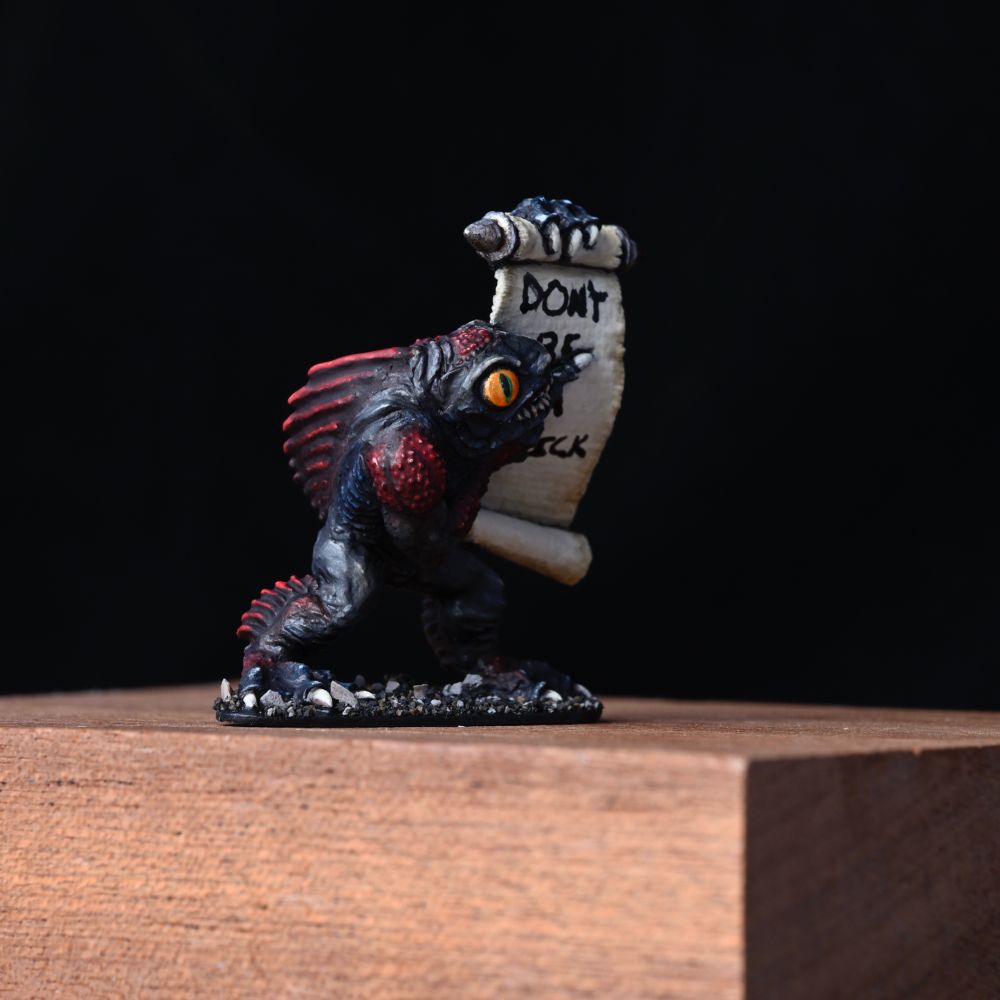  a weird fishy looking guy points to a scroll he holds up. He is black with red bits and bright orange eyes. He also has some dark blue hints around the gills. His scroll states some good advice for life: Don't be a dick.