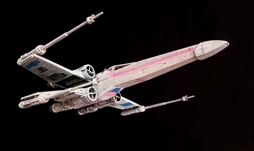 An X-Wing painted in Trans Pride colours, seen from a low angle, from the right side. The X-wing is on a clear black background. It is mostly white, with wing markings in pale blue and a long pink stripe down the sides of the body from the nose. There is another repeated pattern of blue and pink on the underside.