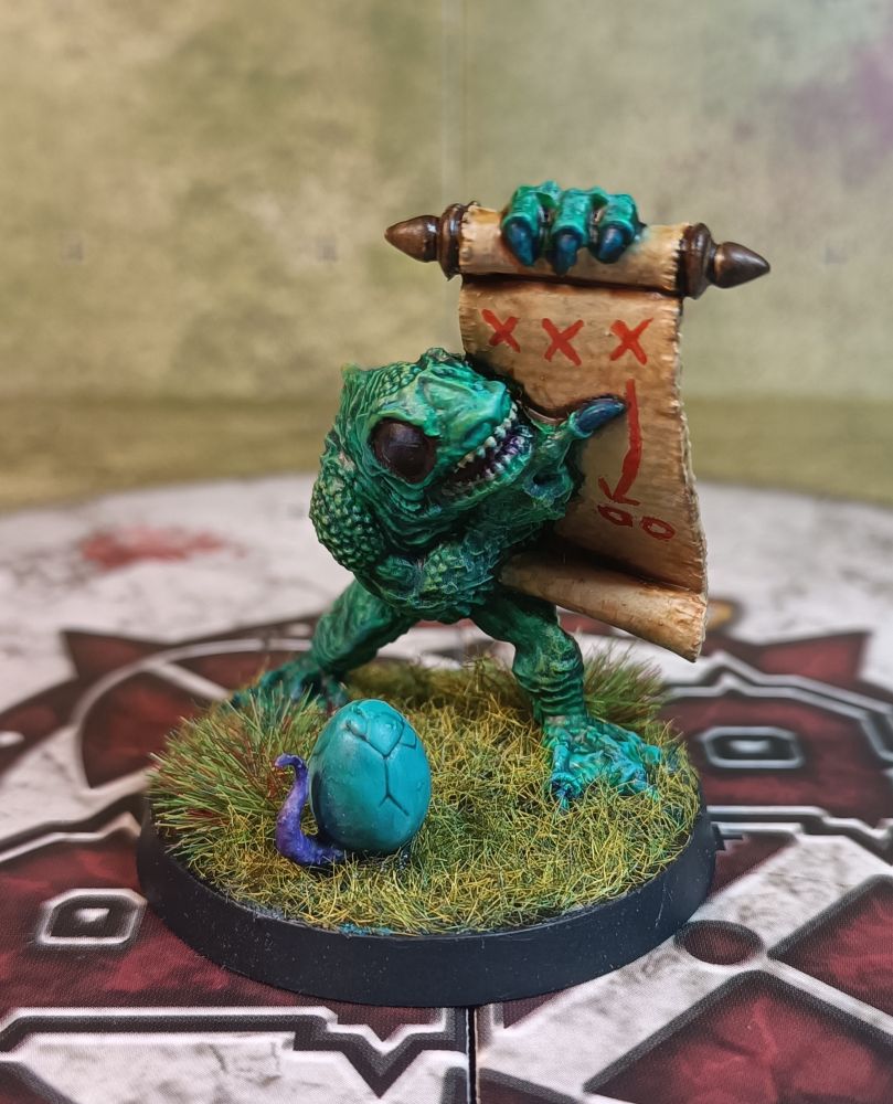Damo the Deep One. No one knows where he comes from or how he became a BloodBowl coach but his tactical genius on the pitch cannot be denied. He resembles a fish-man, his scales/skin is blueish-green and his eyes are large and black. He is holding a large paper scroll with a match-winning play on it in red ink.
