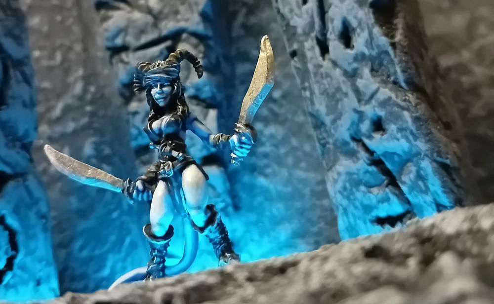 Keynsey the Tiefling Rogue. A clearly sexy little hoofed blue Tiefling with black horns, a rather tight yet revealing black top, and her long black hair gathered in one of those headwraps so beloved by late-80s glam rockers. She is seen from a low angle, giving a bit of a cheeky wink as she wields her two swords. She is stood between three grey stones carved with eldritch symbols, and everything is underlit by a bright blue magical glow.
