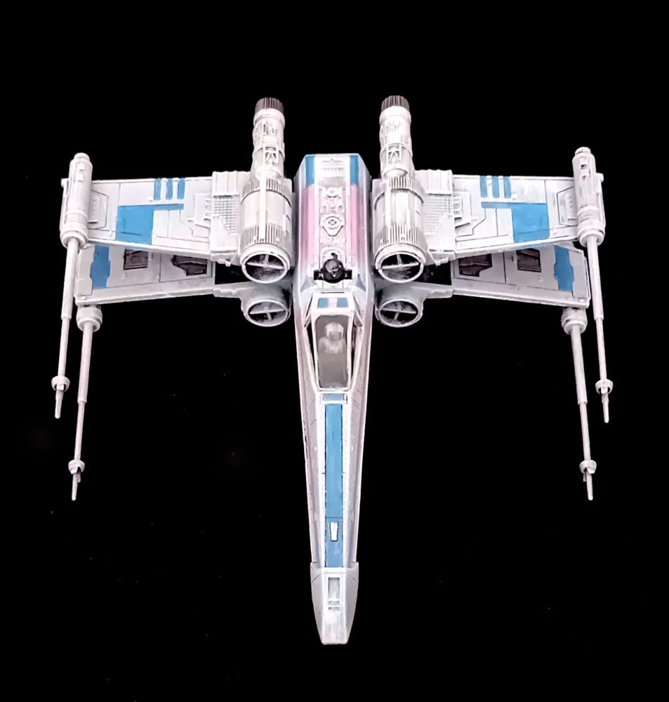 An X-Wing painted in Trans Pride colours seen from above. It is pointing straight downward, still on a black background. The trans flag colours are clearly visible, blue on the wings and the body, and two sections of pink stripes running the length of the body.