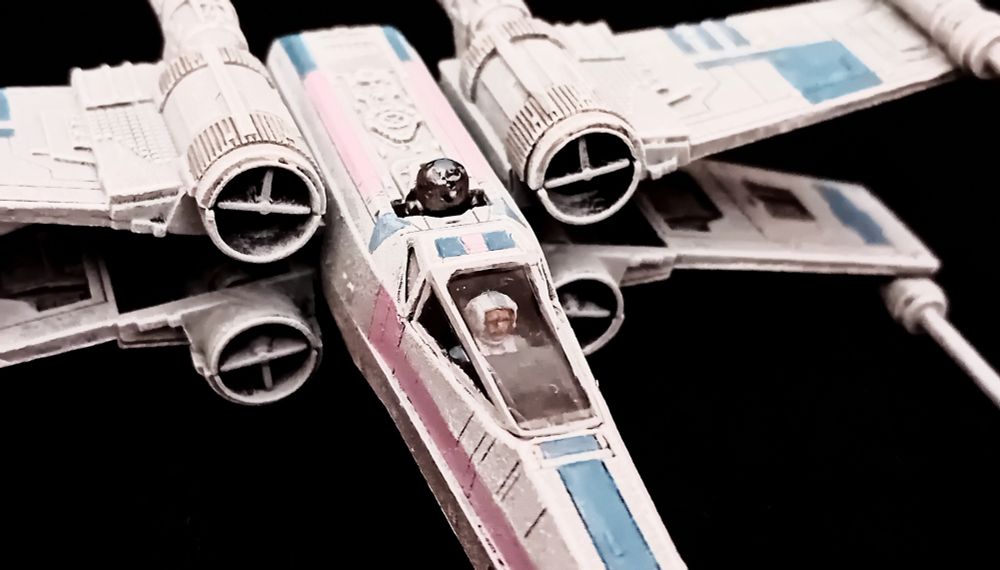 A close-up of an X-Wing painted in Trans Pride colours. The pilot can just be seen, though their blue and pink pilot suit is barely visible. Their astromech droid shines like steel.
