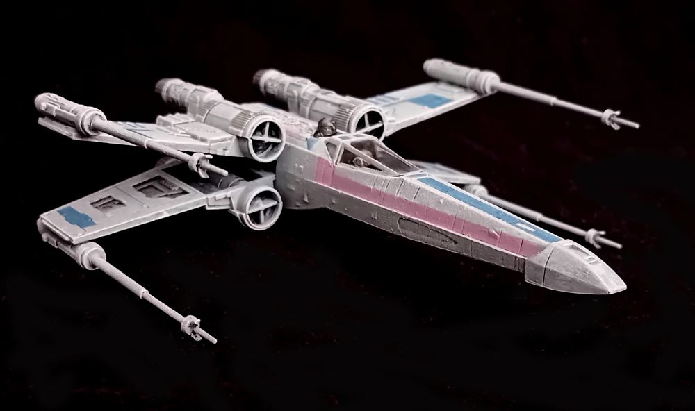 An X-Wing painted in Trans Pride colours, seen from the front right quarter. The wing colours are more visible and a blue stripe can be seen running down the top of the body from the nose.
