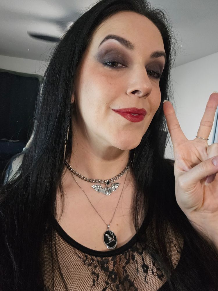 Selfie of me. I'm a pale lady with long dark hair. I'm wearing some smoky eye makeup, dark red lips, some silver bat and raven jewelry, and you can see the top of my Black lace dress. I'm smiling slightly with raised eyebrows and flashing a peace sign. 