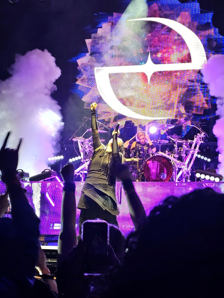Amy Lee wirh purple and smoke all around her, mic to her mouth as she looks up with her fist in the air. You can see silhouettes of crowd members in the foreground with their hands in the air.