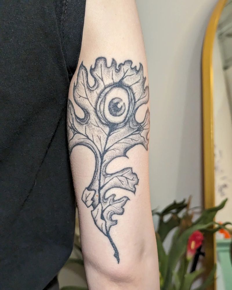 An oak leaf with an eyeball at the top main part of the leaf. Black and grey with stipple shading, located on the back of the arm 