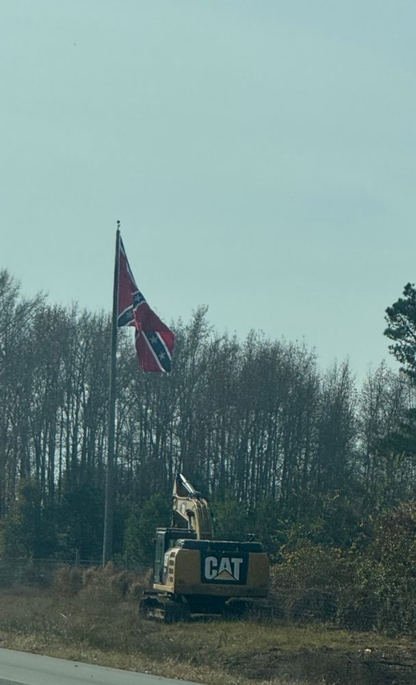 The Confederate Flag on the side of a highway blowing in the wind. There is a CAT construction vehicle off to the side 