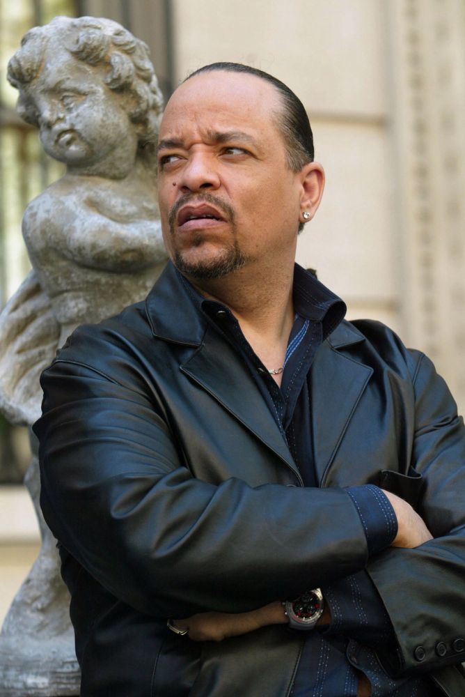 Ice-T looks to the right while standing in front of a statue mimicking its pose of having both arms crossed over each. 