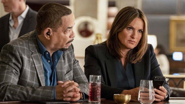 Tutuola sits hands clasped together to the left Benson who is on her phone looking up at someone across the table 