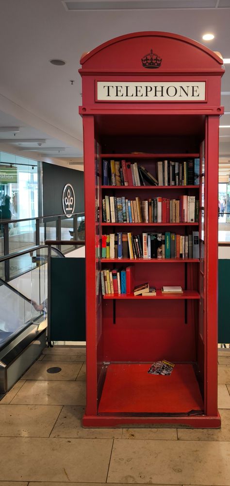 Auf dem Bild ist eine alte rote Telefonzelle zu sehen, wie man sie typischerweise aus Großbritannien kennt. Allerdings wird sie hier nicht mehr als Telefonzelle genutzt, sondern als kleines öffentliches Bücherregal. Im Inneren befinden sich mehrere Regalbretter, die mit einer Vielzahl von Büchern gefüllt sind – teilweise hochkant gestellt, teilweise gestapelt. Unten im Regal liegen außerdem ein paar Zeitschriften oder Hefte lose auf dem Boden der Zelle. Oben an der Zelle ist ein Schild mit der Aufschrift 