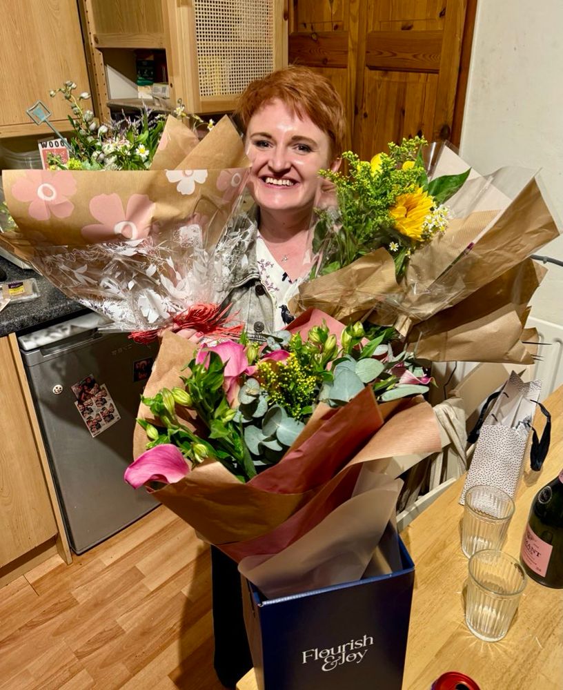 Laura grins in a kitchen, her arms filled with lots of bouquets of flowers and her head just poking above them