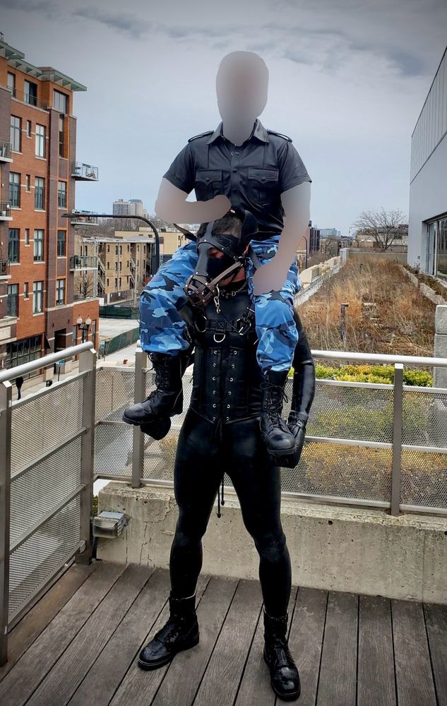 A person in a full black silicone bodysuit is styled as a human horse, complete with a sleek leather corset featuring prominent lace-up details. The figure stands tall in glossy black boots on wooden decking, embodying the aesthetic of human pony play.

Mounted atop this impressive pony sits a rider wearing a military-inspired black shirt with epaulets and shoulder details, paired with distinctive blue camouflage cargo pants. The contrast between the structured military-style top and the tactical camo bottoms creates a commanding presence. The rider's combat boots rest in the pony’s hoofs, which are wrapped in leather puppy mitts that reflect the pony’s hybrid identity as a ponyhound.

The scene unfolds against a dramatic urban backdrop of brick apartment buildings and city architecture. The overcast sky casts a moody atmosphere over the rooftop setting, where metal railings frame the edge of the balcony. In the background, patches of early spring vegetation can be seen on an adjacent rooftop garden area, adding a touch of nature to the metropolitan environment.

This powerful image captures the intersection of alternative lifestyle expression and urban living, showcasing the artistry and dedication involved in human pony play relationships. The composition balances intimate personal expression with the expansive cityscape beyond, creating a compelling visual narrative about community, identity, and connection.

#HumanPonyPlay #LeatherCommunity #AlternativeLifestyle #UrbanScene #SiliconeFetish #KinkPositive #PetPlay