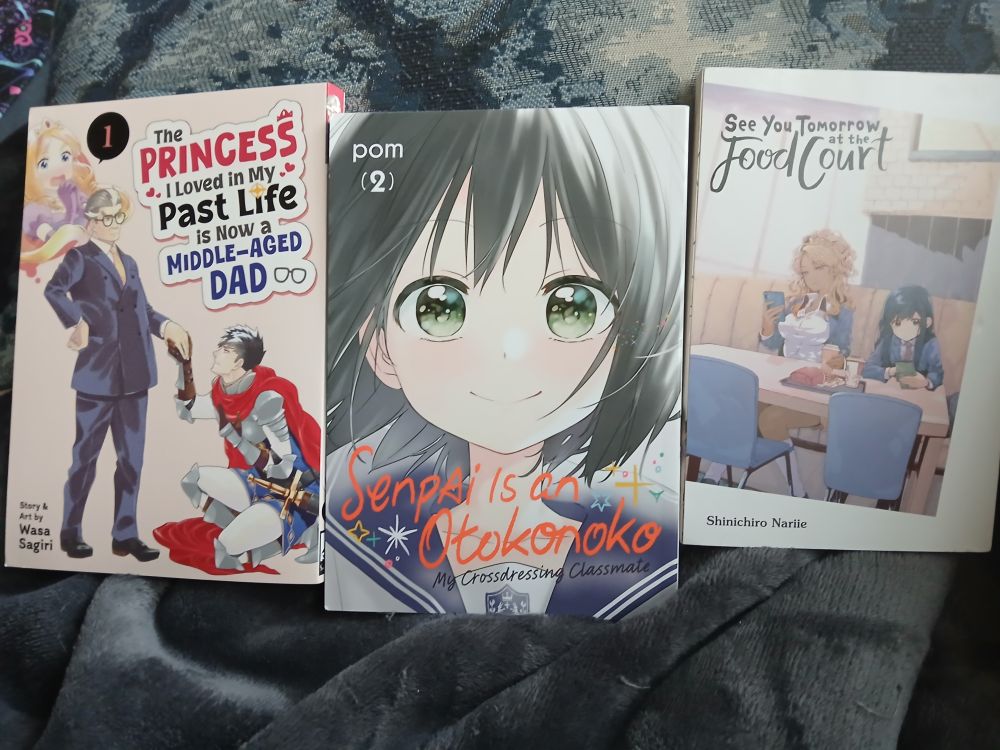 A photo of three volumes of manga. From left to right, they are The Princess I Loved in My Past Life is Now a Middle-Aged Dad volume 1 by Wasa Sagiri, Senpai is an Otokonoko volume 2 by Pom, and See You Tomorrow at the Food Court by Shinichiro Nariie.