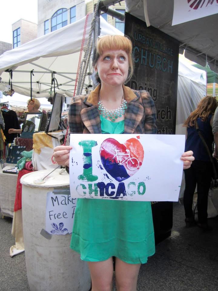 woman in short flowy teal-green dress with matching chunky necklace. Wears plaid riding style jacket over top. Holds homemade print that says "I heart Chicago."