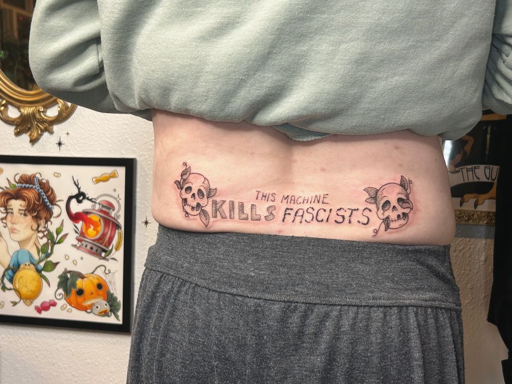 [ lakesidettt ]

A black and gray lower back tattoo with the quote “this machine kills fascists” with two asymmetrical cartoon skulls with cracks and broken teeth. Vines are growing out of the skulls. 