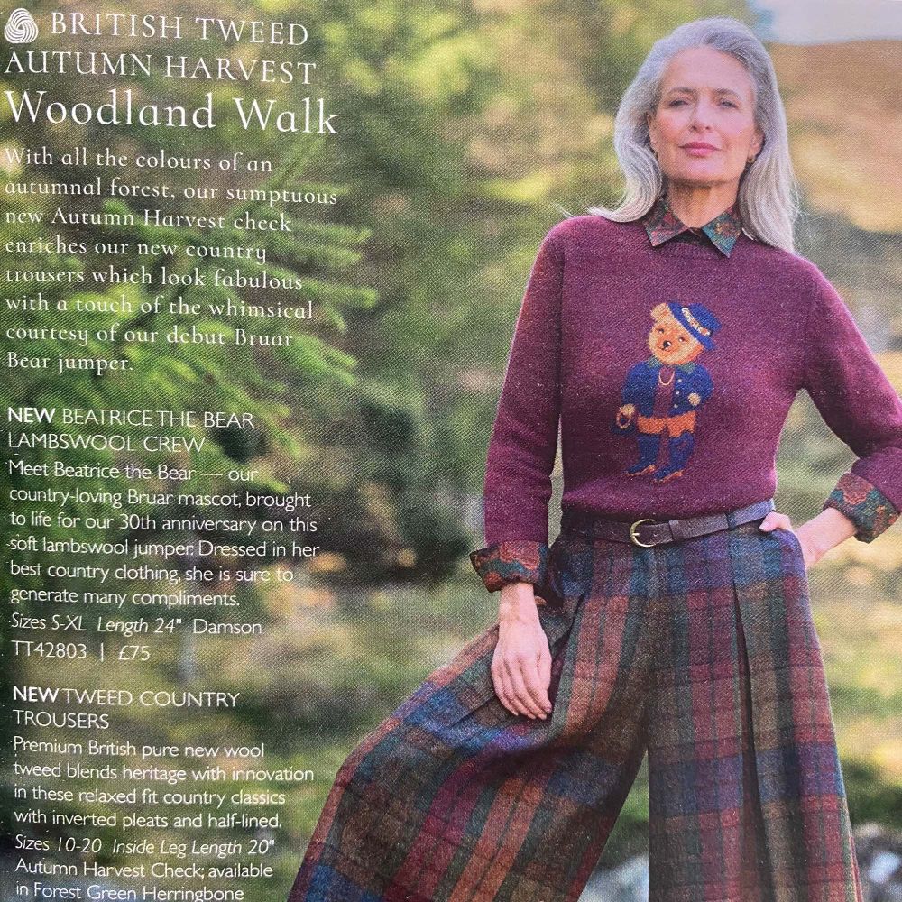 A woman model stands outside wearing outsize tartan 'country' trousers in muted green and purple, and a purple woollen jumper with a cutsie bear cartoon mascot. She looks commandingly into the distance.