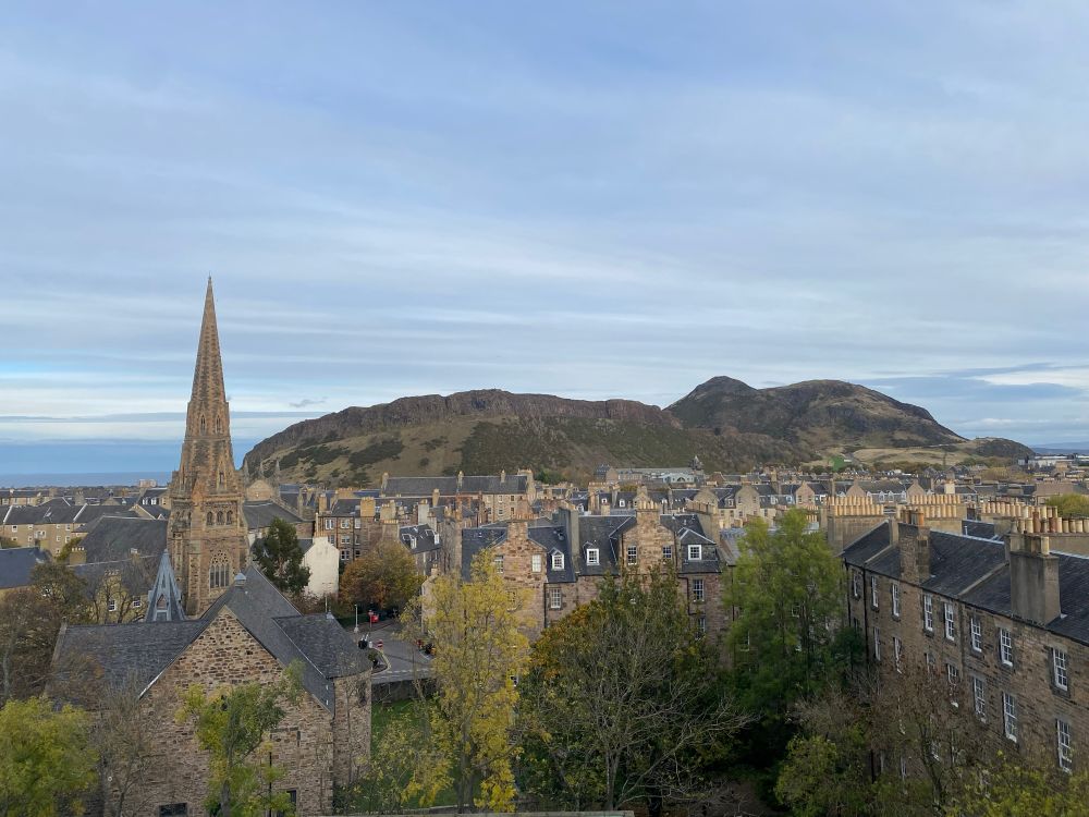 View east over Edinburgh across Arthur’s Seat, autumnal leaves, grey sky, and the gothic rocket spire of Buccleugh Free Church. 