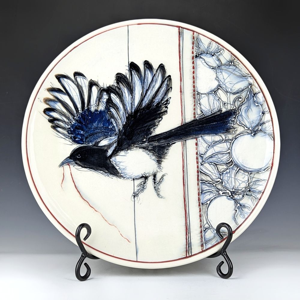 Ceramic platter featuring a magpie with a red thread in its beak while flying away from a tapestry pattern of apples and leaves.