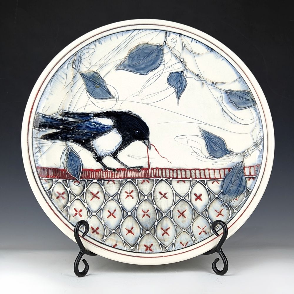 A ceramic platter depicting a magpie perched upon a stitched pattern. The magpie is pulling on a thread from the stitching. The background is made up of blue leaves and branches.