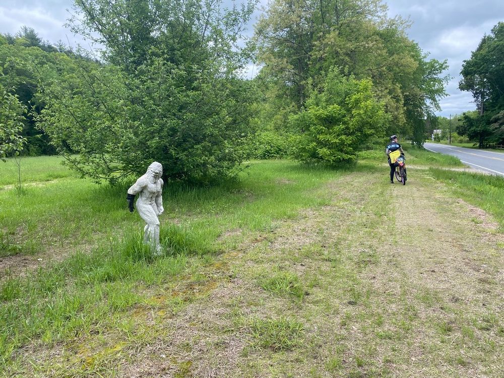 A cyclist wearing a black HUP kit jersey, yellow and grey jacket, black pants, and riding a yellow bike with purple rims is riding on an off-road route of field and grass adjacent to a road. There is a sculpture of a Big Foot, roughly 5 feet tall and made of what looks like concrete off to the left of the trail, and someone has put a black glove on one hand. This is on part of an “Inactive Railroad Road” (Ashuelot Recreational Rail Trail) starting in Ashuelot, NH and continuing to Hinsdale, NH, along the border of NH and VT.