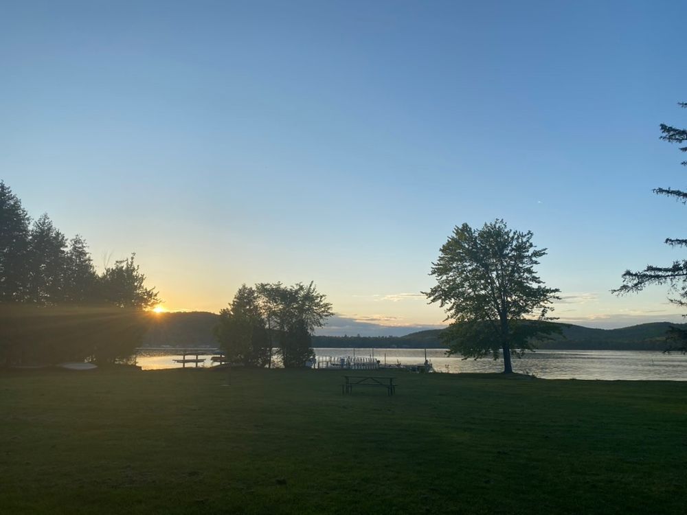 A picture of a sunset at a lake. There is a grassy field that leads up to a dock with a couple of boats and a person sitting with their feet in the water, and there are a few trees along the perimeter of the lake. The sun is kissing the top of a mountain top in the distance, and other mountains are visible as well. The sky is clear except at the horizon where there are some thin clouds reflecting the sunlight. Light rays are streaming from the sun.