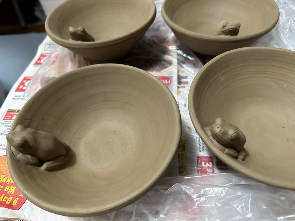 Four greenware bowls arranged in a square in a paper covered table. A little sculpture frog sits inside each peaking over the lip.