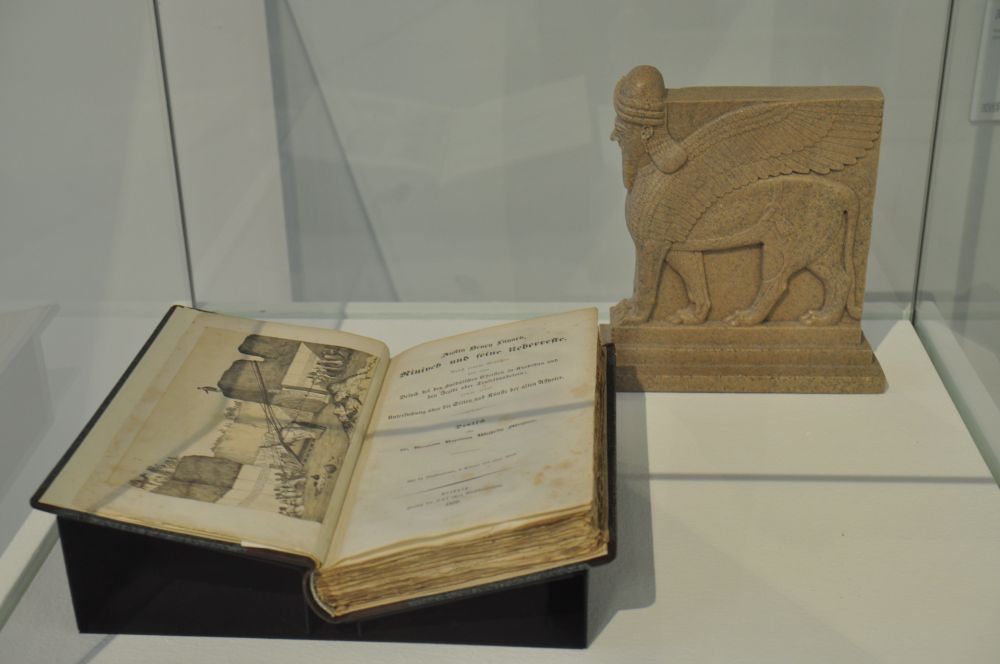Das Bild zeigt einE Vitriene mit einem aufgeschlagenem Buch (Austin Henry Layard:  Niniveh und seine Ueberreste) und dem Modell eines Lamassu.