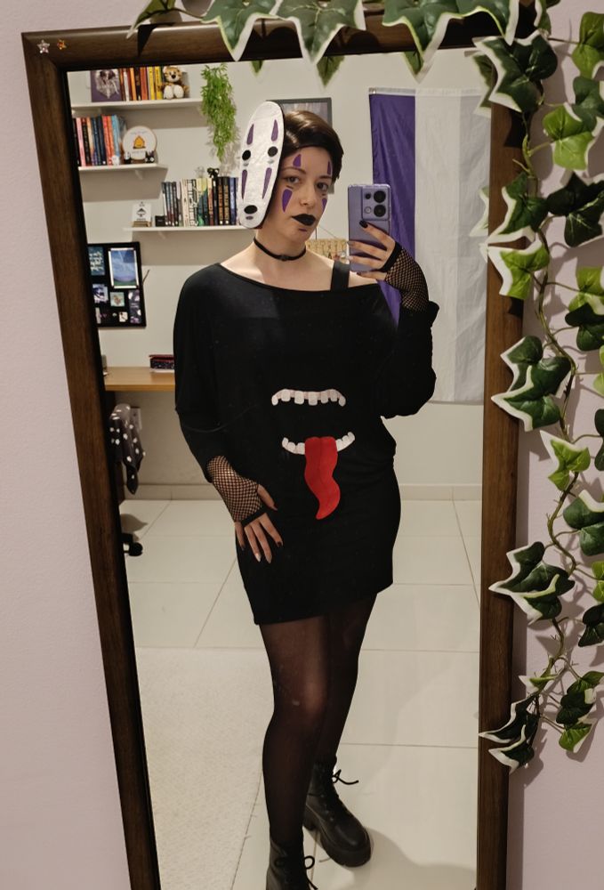 Foto minha na frente do espelho, estou vestindo um vestido preto largo na parte de cima, no meio dele tem uma boca aberta com dentes e uma língua vermelha esticada pra fora, estou usando uma máscara do personagem kaonashi na cabeça, ele tem o rosto branco com marcas vesti cais roxas, olhos e boca pretos. No rosto fiz uma maquiagem com as mesmas marcas do personagem e com batom preto. Estou vestindo uma meia calça fumê e coturnos. Sou uma mulher branca de 28 anos, cabelo castanho curto na altura da orelha. 