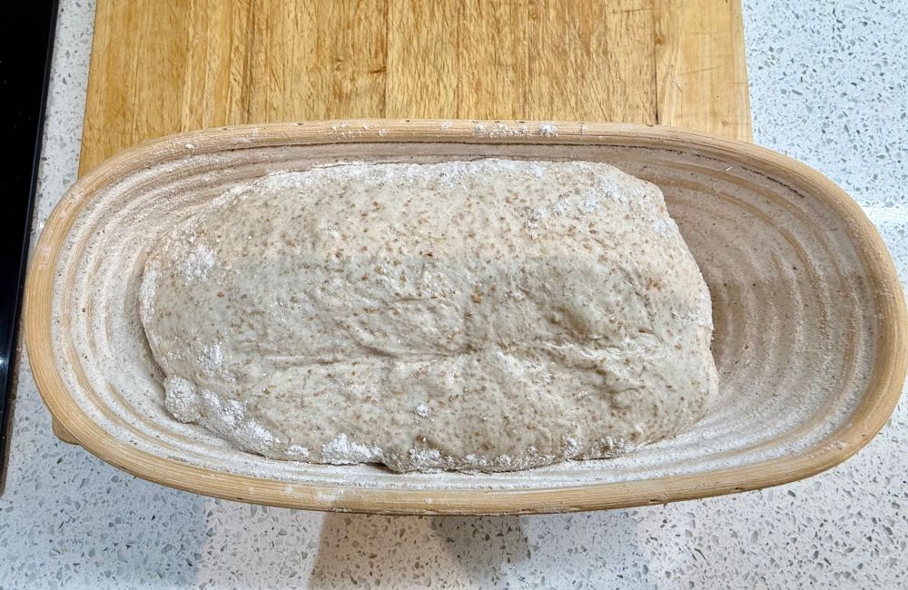 Bread dough in an floured oval cane banneton. Pictured horizontally, it sits on a wooden chopping board which is on a white surface speckled in grey