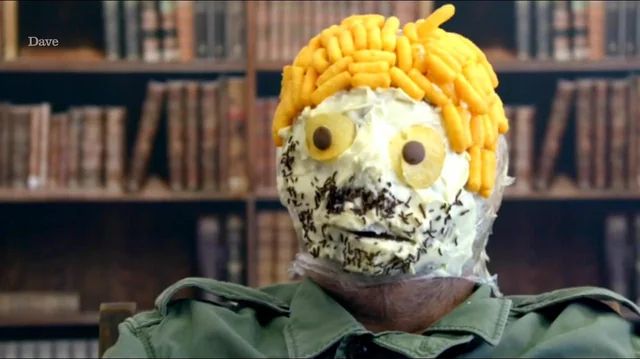 Still of the first Taskmaster: Champion of Champions special, depicting Bob Mortimer wearing his edible mask. The mask is made of icing, chocolate sprinkles, Wotsits, and potato crisps, and affixed to Bob’s face with cling film.