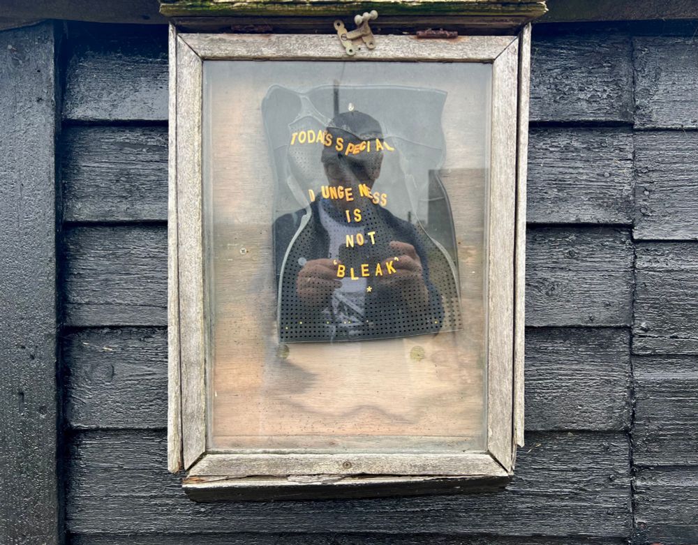 Reflection of photographer in glazed box frame containing a sun-warped plastic sign that reads ‘ TODAYS SPECIAL - DUNGENESS IS NOT “BLEAK”  * ‘