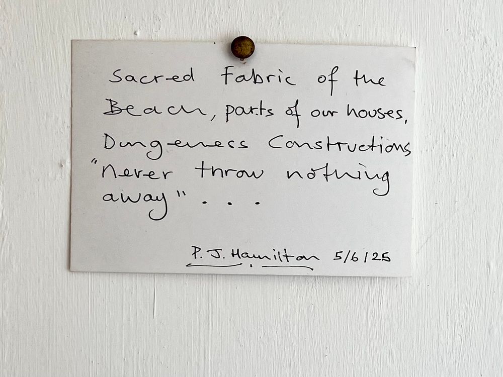 Handwritten note on white card pinned to the wall reads ‘Sacred fabric of the Beach, parts of our houses, Dungeness Constructions “never throw nothing away”… 
P. J. Hamilton 5/6/25’
