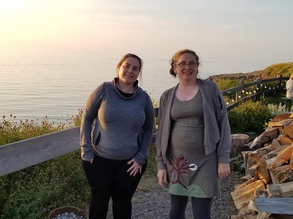 Alison Etter (brown hair, black trousers, grey sweater) and me (blonde hair, green dress with tree motif) on the cliff over the Atlantic in Chéticamp, Cape Breton Island, at sunset, August 2021.